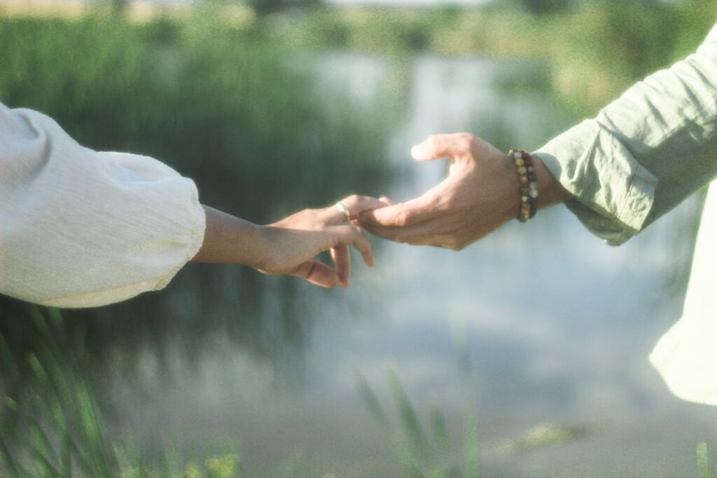 A couple gently holding hands near a calm body of water on a sunny summer day.